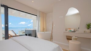 A bedroom with a large bed, a bathtub, a vanity with a mirror, and a view of the sea.