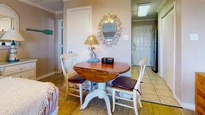 Habitación pequeña con una mesa de madera, dos sillas, una lámpara, un espejo y una cama con manta a rayas.