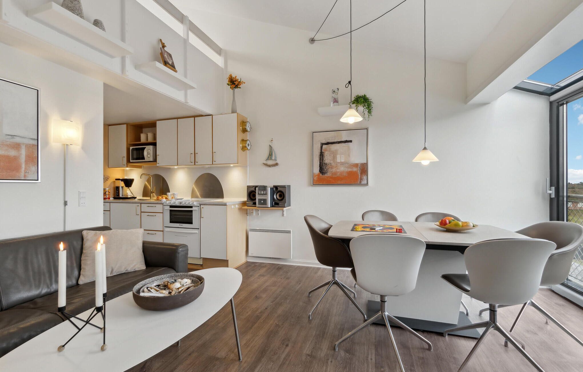 Una cocina moderna con armarios blancos, un comedor con mesa y sillas blancas, y una zona de estar acogedora con sofá y velas.