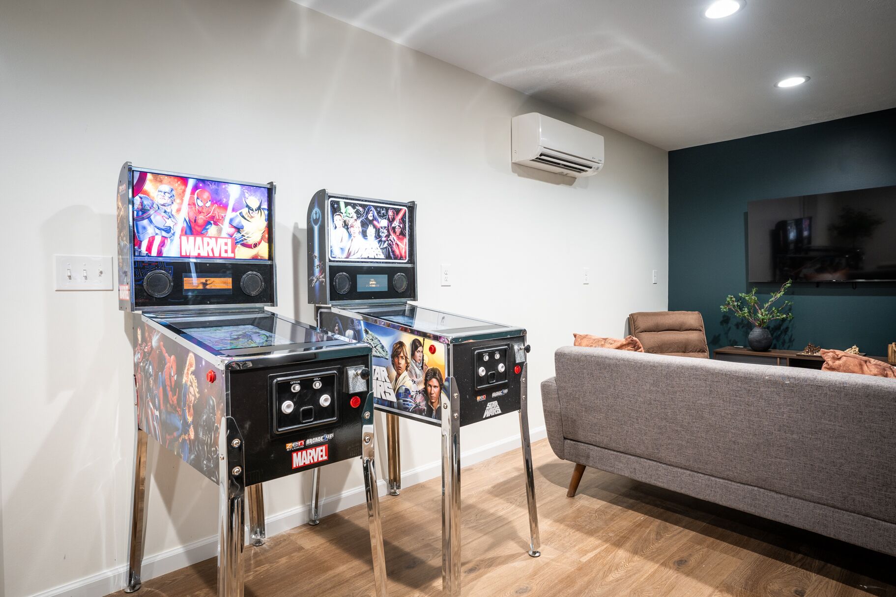 Två arkadspel med teman från Marvel och Star Wars, en grå soffa och en väggmonterad luftkonditioneringsanläggning.