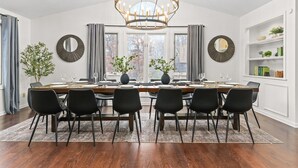 Ein Esszimmer mit einem großen Holztisch, gedeckt für sechs Personen, umgeben von schwarzen Stühlen, einer Kristallleuchtre und zwei großen, mit Vorhängen versehenen Fenstern.