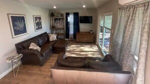 A spacious living room with a dark brown sofa set, a small side table, and a cabinet. There are two framed pictures on the wall, a ceiling light, and an air conditioning unit.