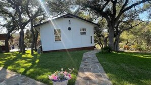 A small white building with a stone pathway leading to it, surrounded by trees and a grassy area.