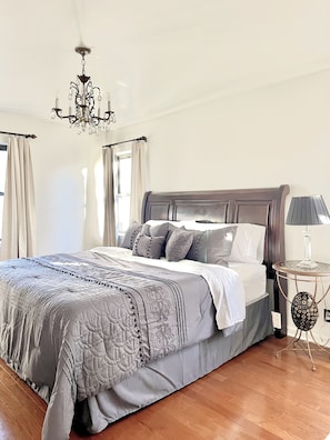 A bedroom with a large bed, a chandelier, a bedside table with a lamp, and curtains on the windows.