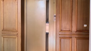 Ein Aufzug mit metallischem Innenraum und Bedienfeld, begleitet von Holzwandverkleidung, einer Tür links und einer an der Wand montierten Überwachungskamera rechts.