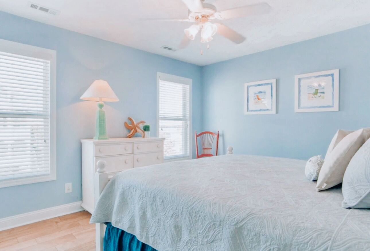 Un dormitorio con cama, cómoda, silla y dos ventanas con persianas.