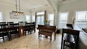 A spacious dining area with a piano, wooden chairs, and a large window offering a view. - “Coastal Haven” Gorgeous 6 bedroom home pool (North Topsail Beach)