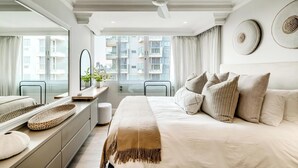 Een moderne slaapkamer met een groot bed, een plafondventilator, een raam met uitzicht op gebouwen en een nachtkastje met een spiegel en een plant.