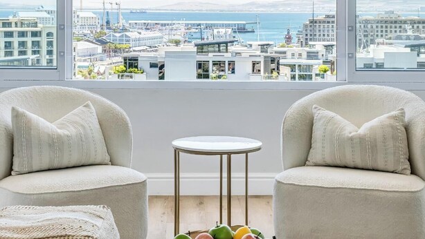 Een moderne woonkamer met twee fauteuils, een klein rond tafeltje en een schaal met fruit. De kamer heeft grote ramen met uitzicht op een stadsbeeld met gebouwen en een waterpartij.