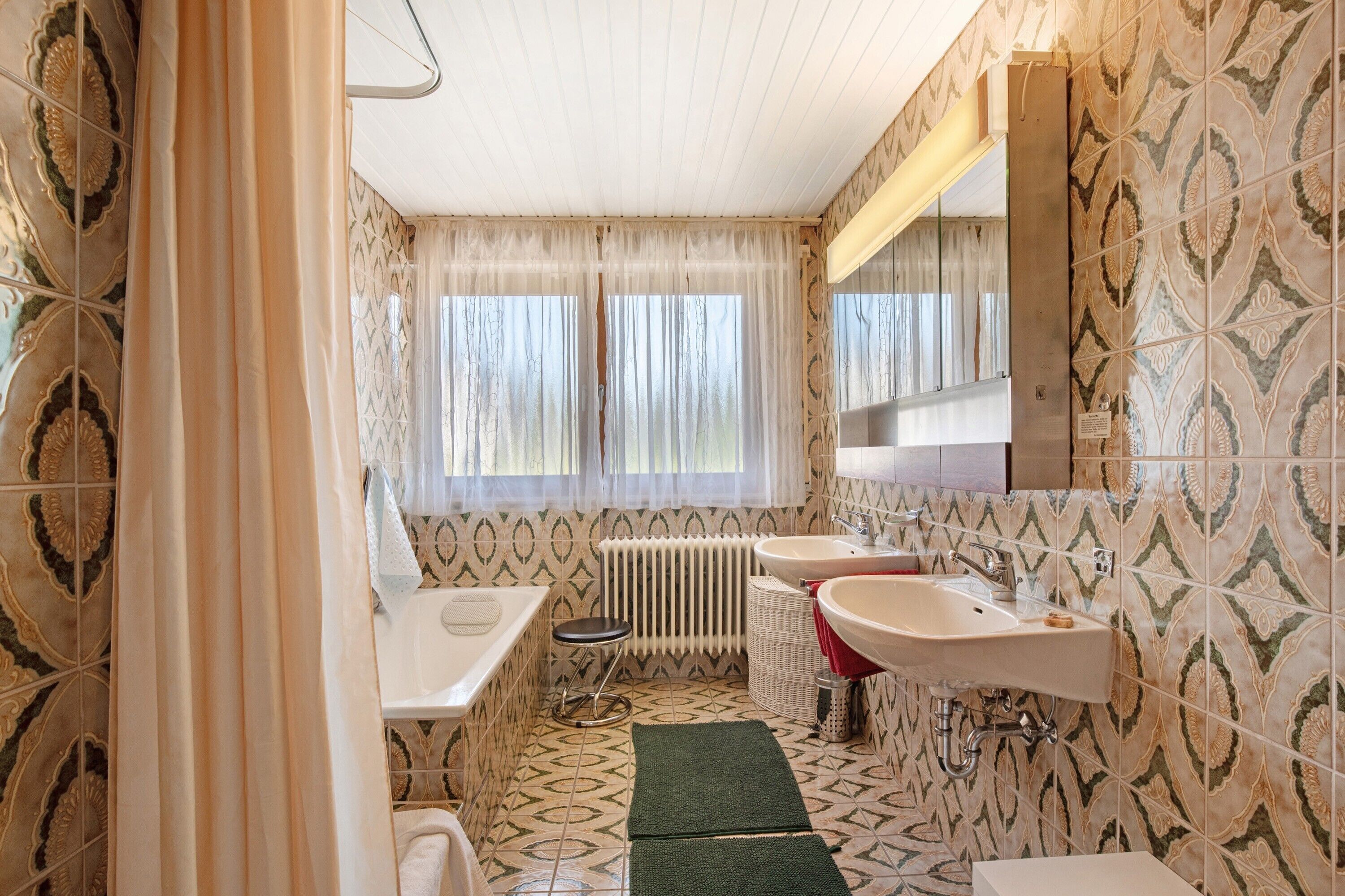 Une salle de bain avec des carreaux décorés, deux lavabos, une baignoire, un radiateur et une fenêtre avec des rideaux.