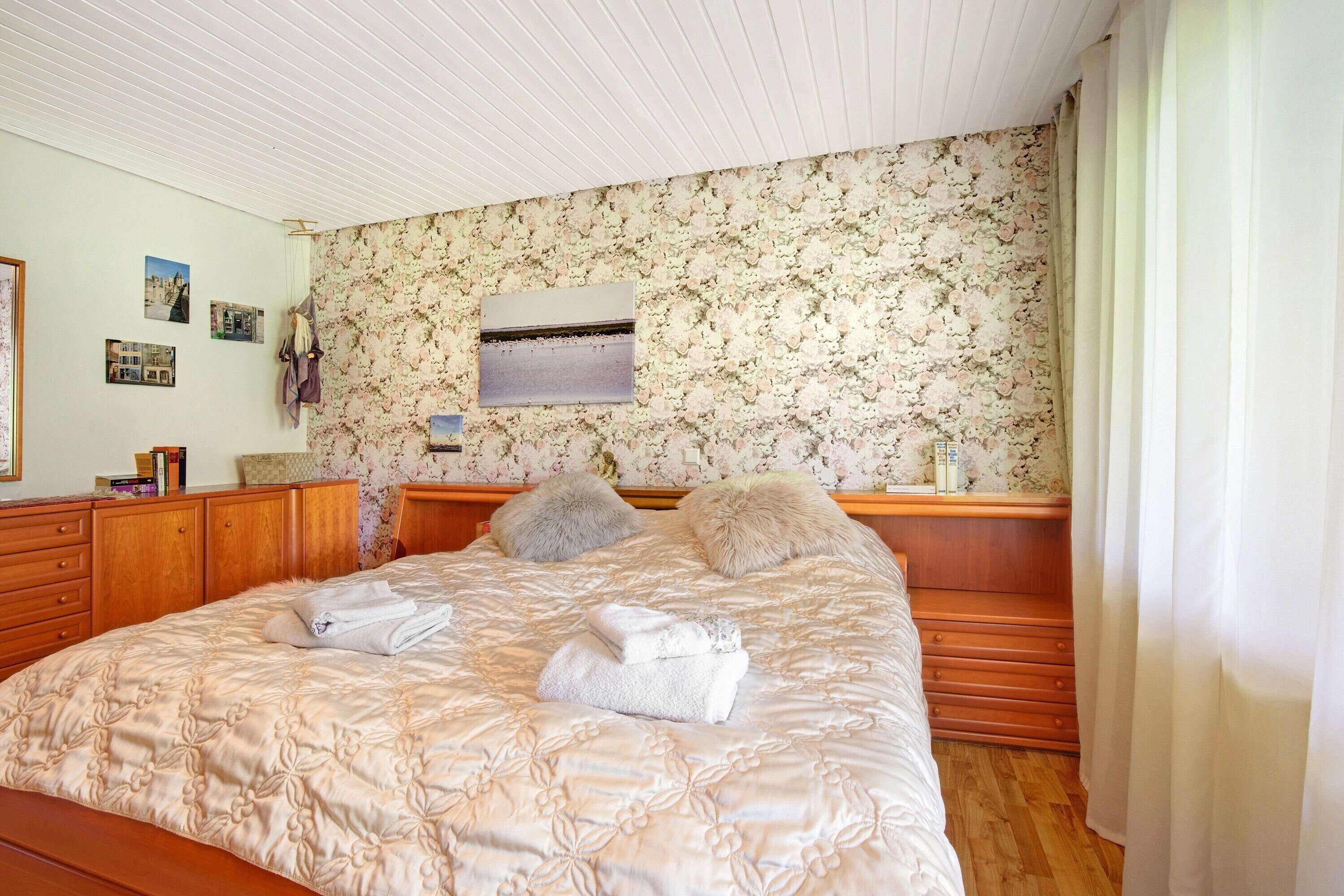 Un dormitorio con papel tapiz floral, un cabecero de madera, una cama con colcha color beige y una mesita de noche con lámpara.