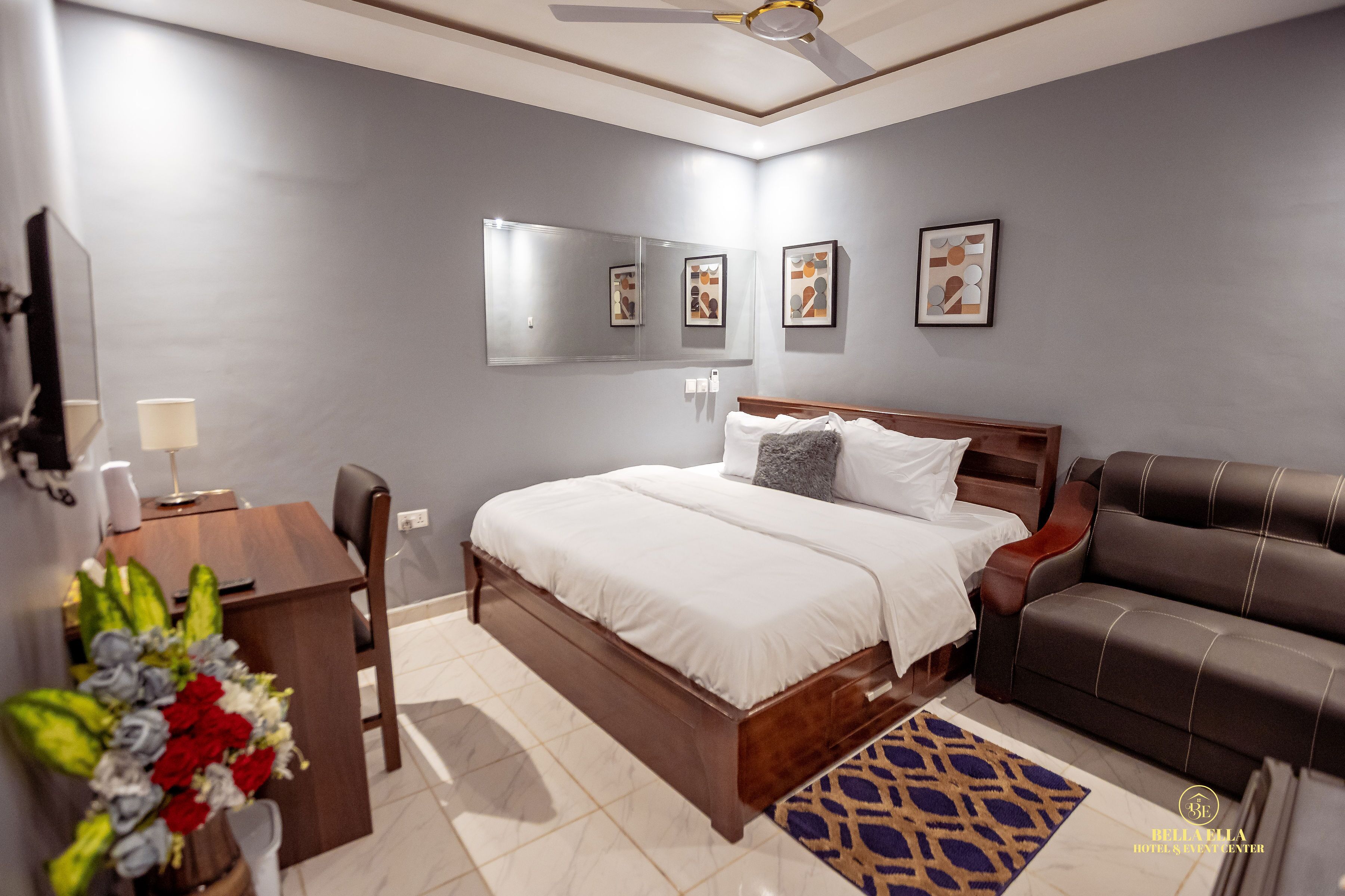 A modern hotel room with a large bed, a desk with a lamp, a chair, a sofa, and a decorative rug.