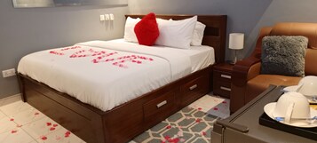 Una cama impecablemente hecha con pétalos de rosa, una mesita de noche de madera con una lámpara, un sillón de cuero marrón y una bandeja con tetera y tazas.