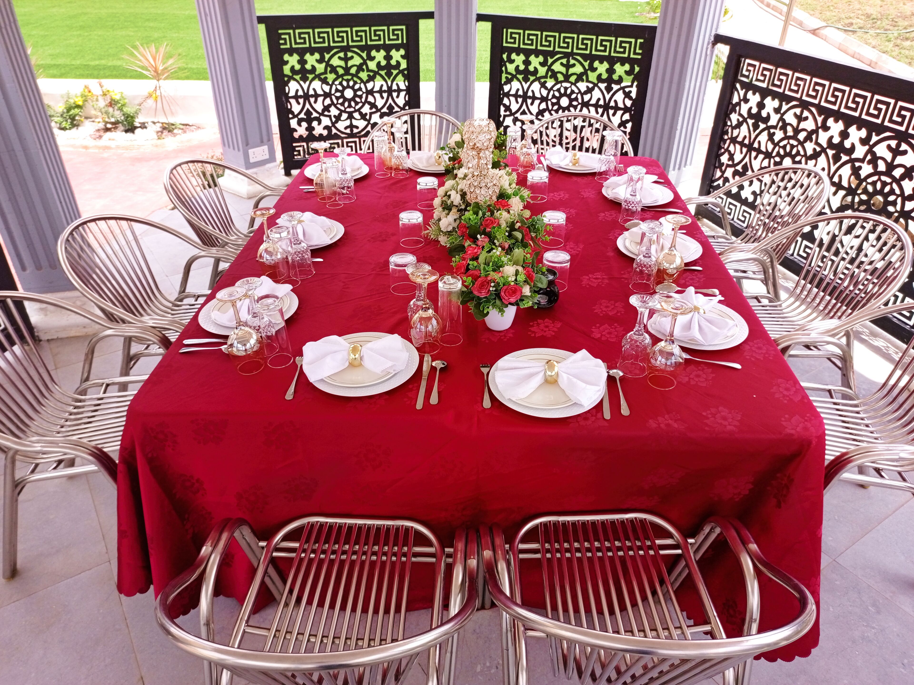 A table set for a formal outdoor dining experience with a red tablecloth, white plates, and silverware, surrounded by metal chairs.