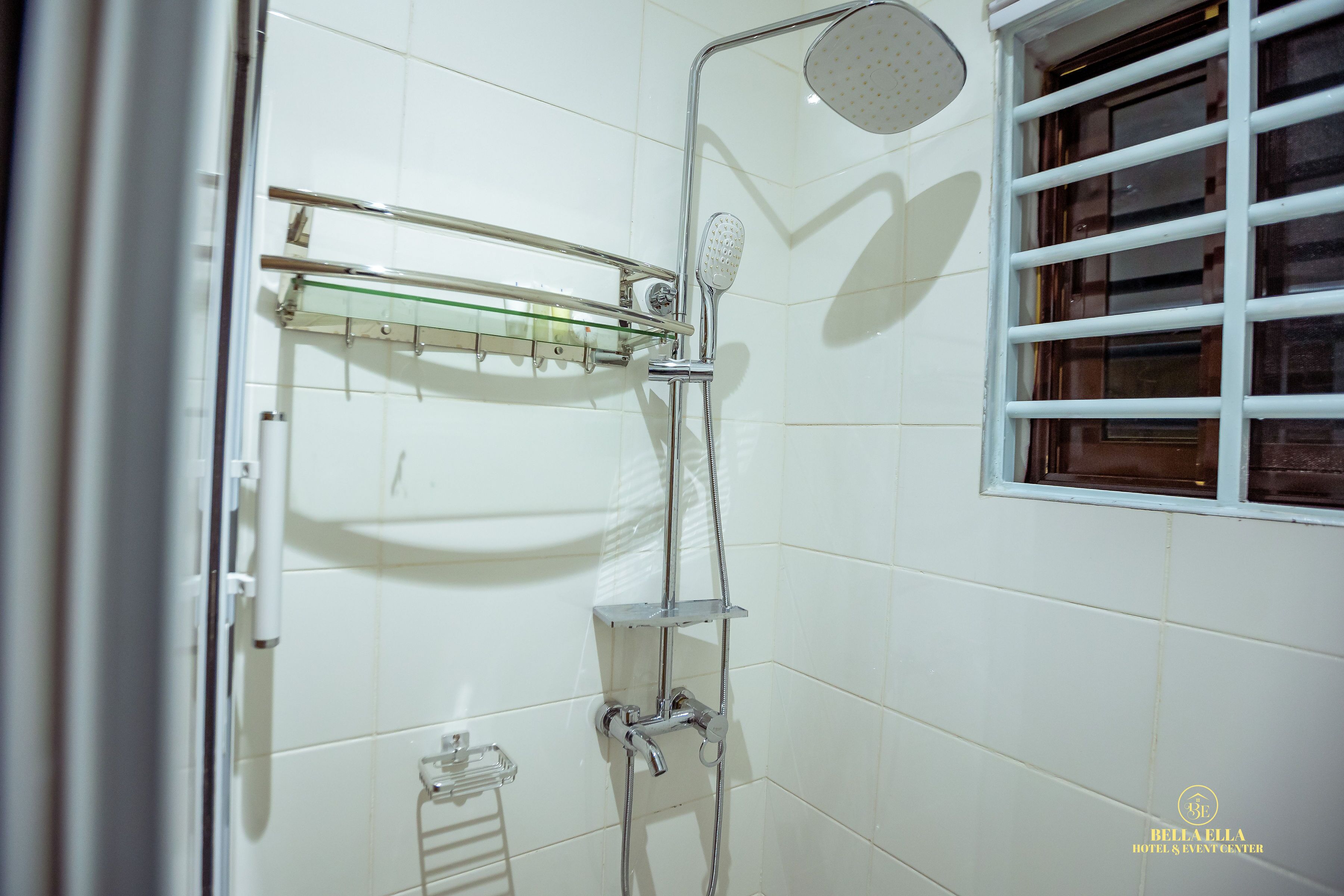 A bathroom with white tiled walls, a glass shower enclosure, a handheld showerhead, and a window with bars.