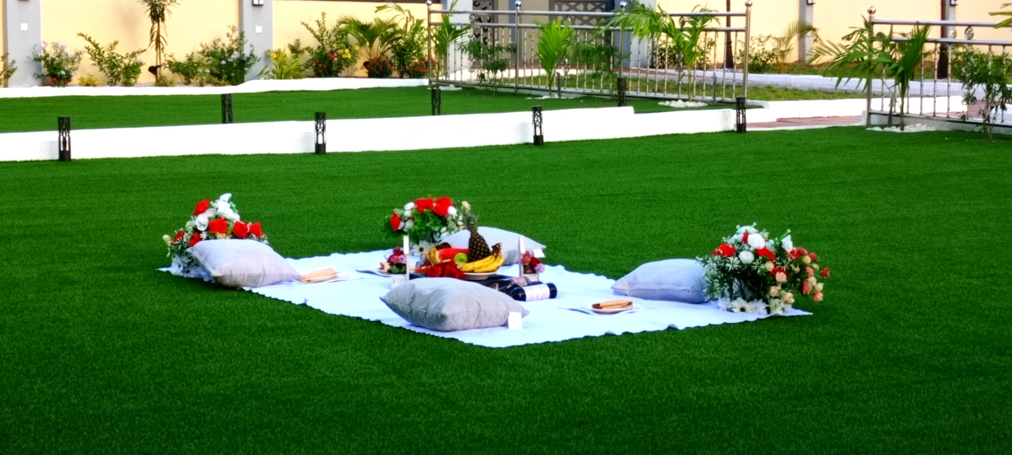 A picnic setup on a grassy area with a white blanket, surrounded by decorative flowers and a bottle.