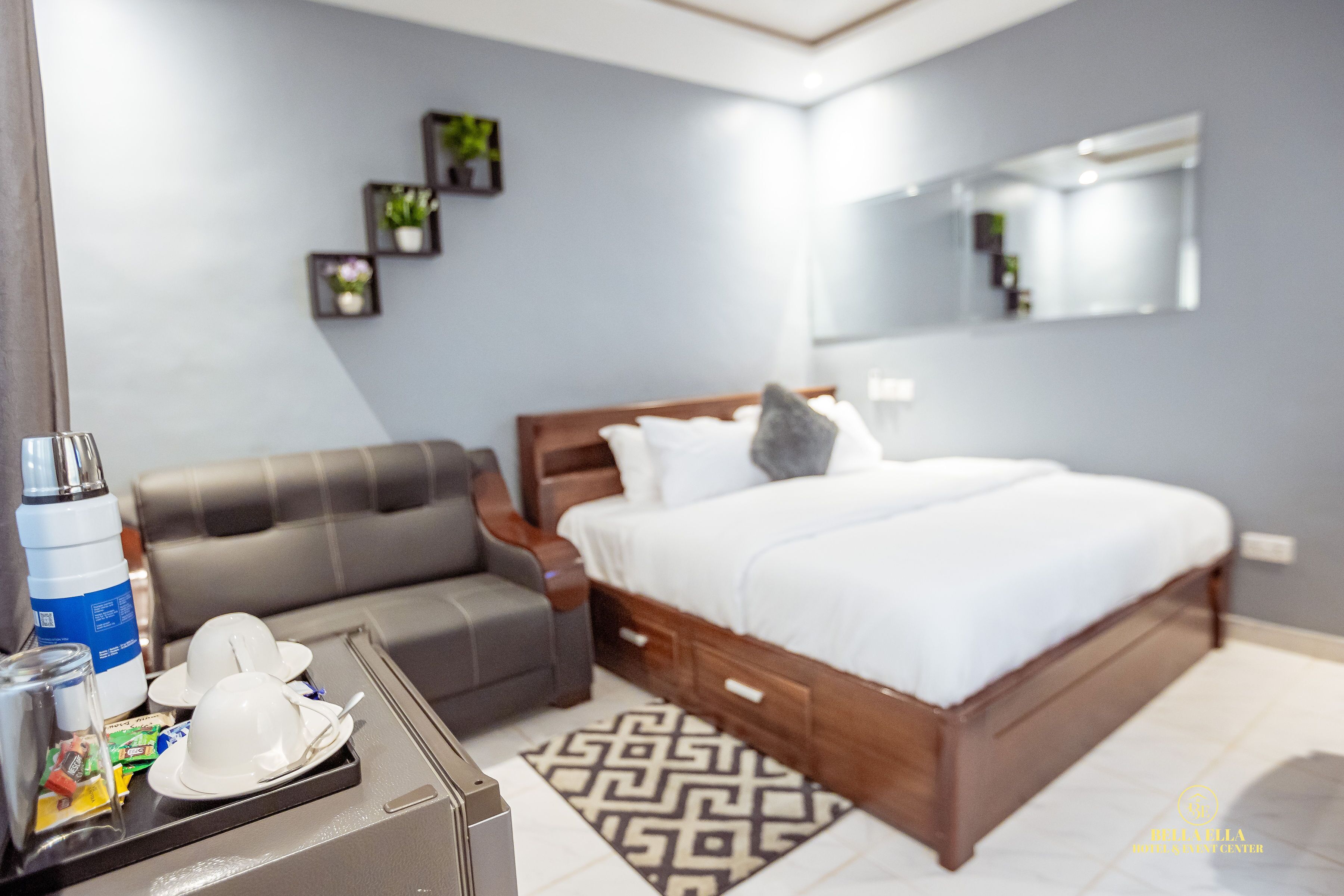 A modern hotel room with a wooden bed, a grey sofa, a small table with amenities, and a geometric patterned rug.