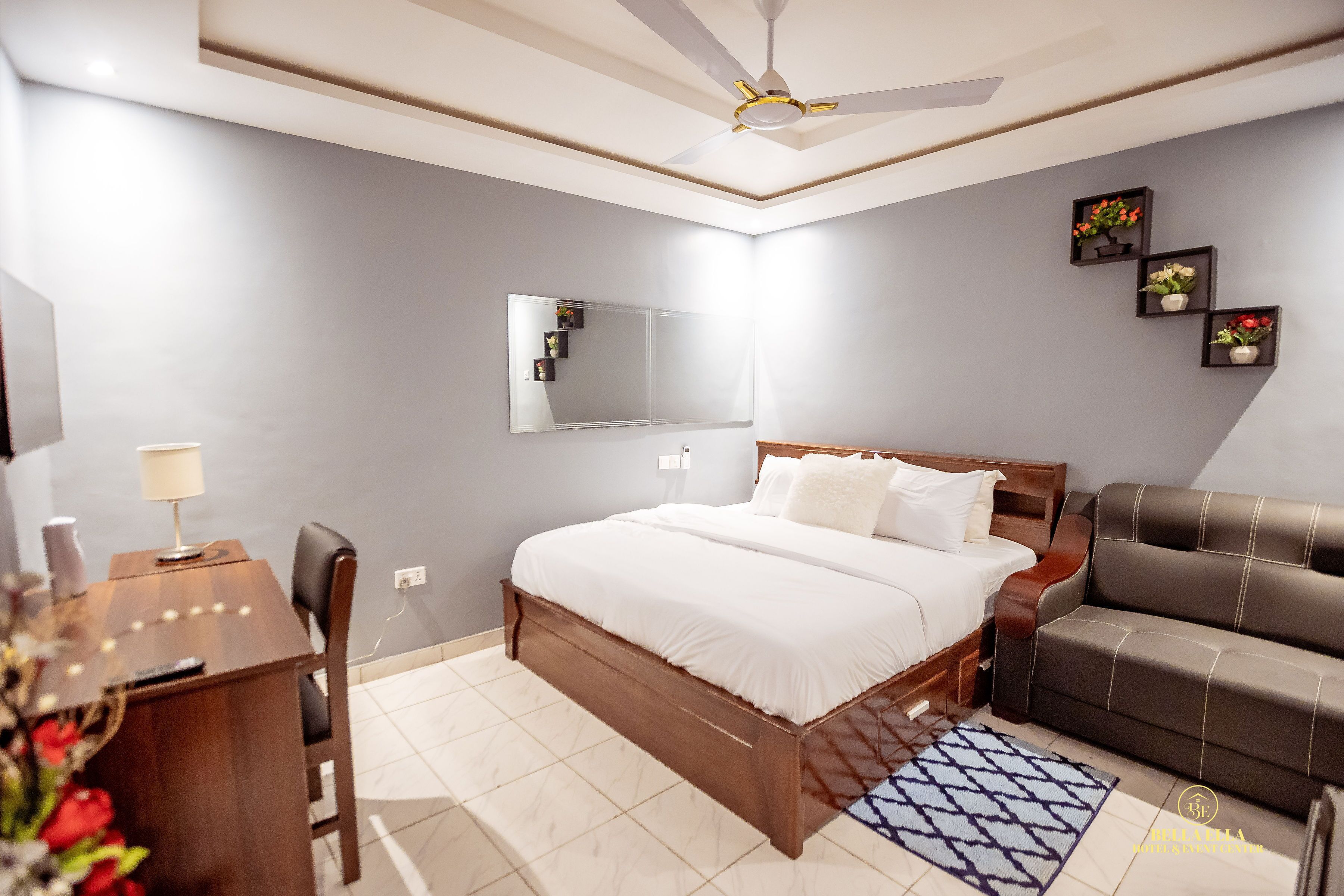 Un dormitorio con cama, escritorio, silla, sofá, ventilador de techo, espejo y estantes en la pared con flores.