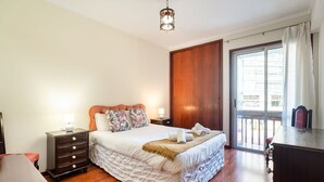 A bedroom with a bed, nightstands, a dresser, a wooden door, and a balcony with curtains. - GuestReady - Varanda de belas lembranças (Funchal)
