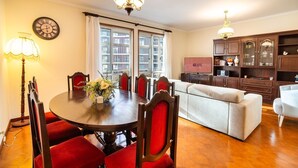 A dining area with a round table, red upholstered chairs, and a vase of flowers. There is a sofa and a television in the background. - GuestReady - Varanda de belas lembranças (Funchal)
