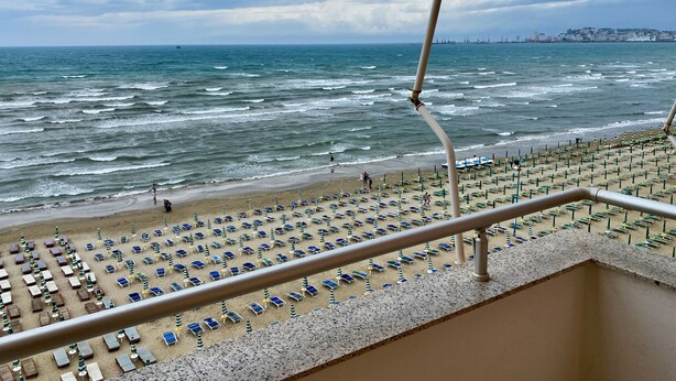 Uma praia com fileiras de espreguiçadeiras e guarda-sóis, um corrimão e vista para o mar e a cidade ao longe.