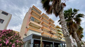 Um hotel com vários andares, varandas, um restaurante com mesas ao ar livre e palmeiras.