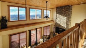 A spacious room with a high ceiling, large windows offering mountain views, a stone fireplace, and a wooden staircase. - New! Peaceful Mountain Views, Deck, Trails nearby, Pool Table. (Mars Hill)