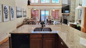 A spacious kitchen with granite countertops, wooden cabinets, and a central island with a sink. There are multiple windows providing natural light and a stone fireplace. - New! Peaceful Mountain Views, Deck, Trails nearby, Pool Table. (Mars Hill)