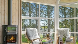 A cozy room with a wood-burning stove, two white furry chairs, a table with a candle and a vase, and large windows offering a view of trees. - "Nayati" - 2km from the sea by Interhome (Saltum)