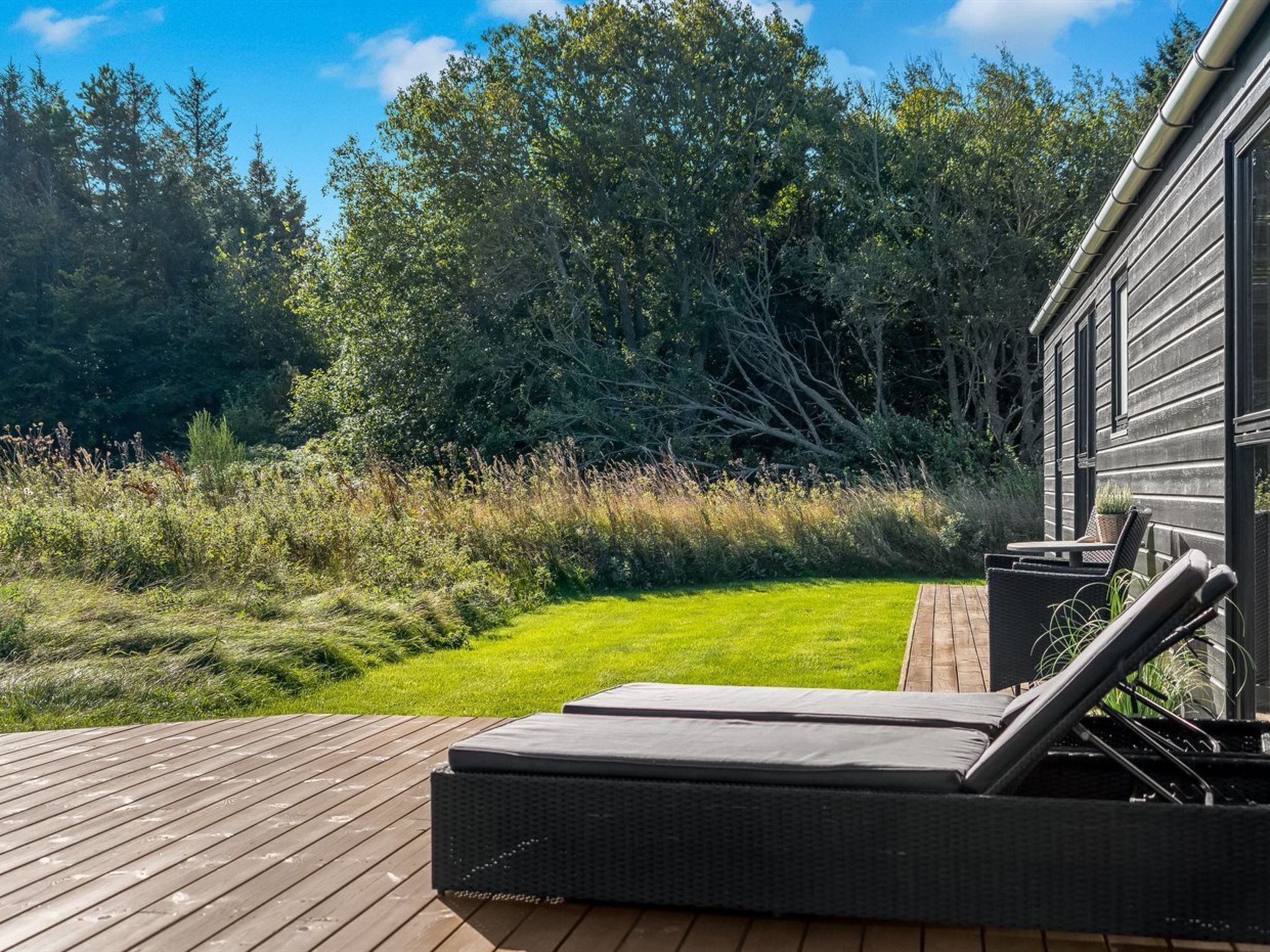 Une terrasse avec un fauteuil de relaxation et une petite table, entourée d’une pelouse verdoyante et de nombreux arbres.