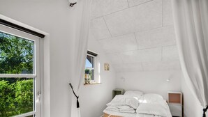 Un dormitorio con un ventanal grande, una cama con ropa de cama blanca, una mesita de noche y un ventanilla con vistas a la vegetación.
