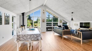 Un loft espacioso con techo alto, una zona de comedor con una mesa larga y sillas, un rincón acogedor con sofá y televisor, y grandes ventanales con vistas a zonas verdes.