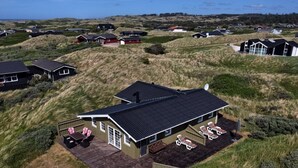 Une vue aérienne d’une maison avec un toit en pente, entourée de dunes herbeuses, et offrant une vue dégagée sur l’océan en arrière-plan.