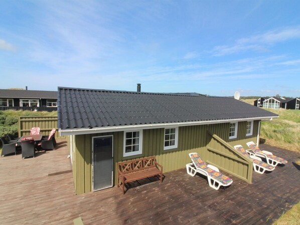 A wooden deck with a house featuring a dark roof, a bench, and sun loungers.