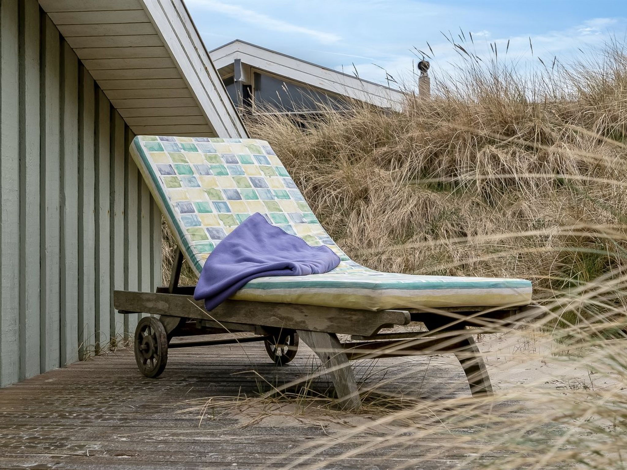 A beach chair with a checkered cushion and a purple towel, positioned on a wooden deck near a building with a sloped roof.