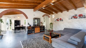 A spacious living area with a grey sofa, wooden ceiling beams, a dining area with a table and chairs, and a kitchen with a built-in cabinet. - Villa Vittorio 6 2 Emma Villas (Imperia)