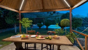 A wooden table set for a meal with various food items and a magazine, surrounded by potted plants and a pool in the background. - Villa Vittorio 6 2 Emma Villas (Imperia)