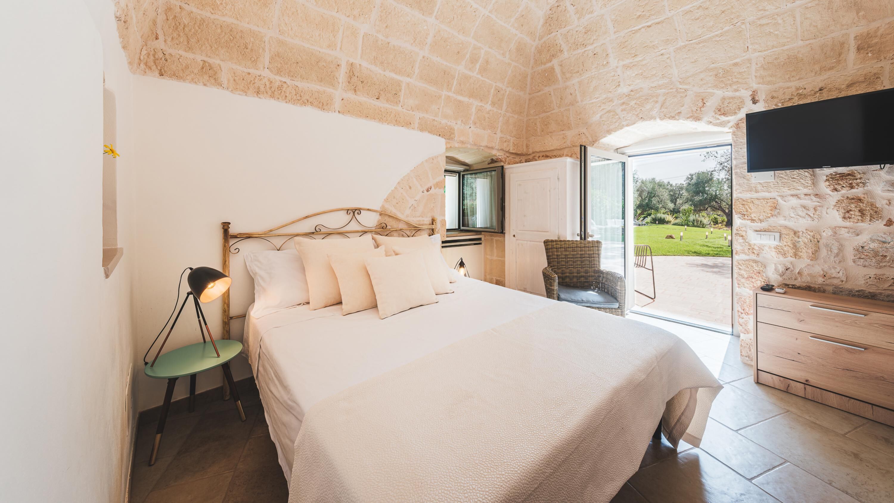 Un dormitorio con pared de piedra, una cama con ropa blanca, una silla de mimbre y un televisor montado en la pared.