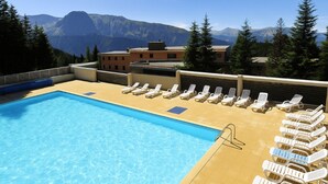 Ein Pool mit klarem, blauem Wasser, umgeben von weißen Liegestühlen und Blick auf die Berge im Hintergrund.