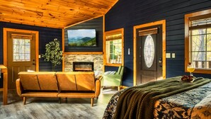 Um quarto aconchegante com teto de madeira, lareira de pedra, um sofá, uma cama com manta verde e uma televisão grande na parede.