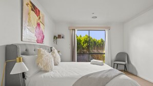 Een slaapkamer met een groot bed, een nachtkastje met een lamp, een stoel, een raam met uitzicht op bomen en een schilderij aan de muur.