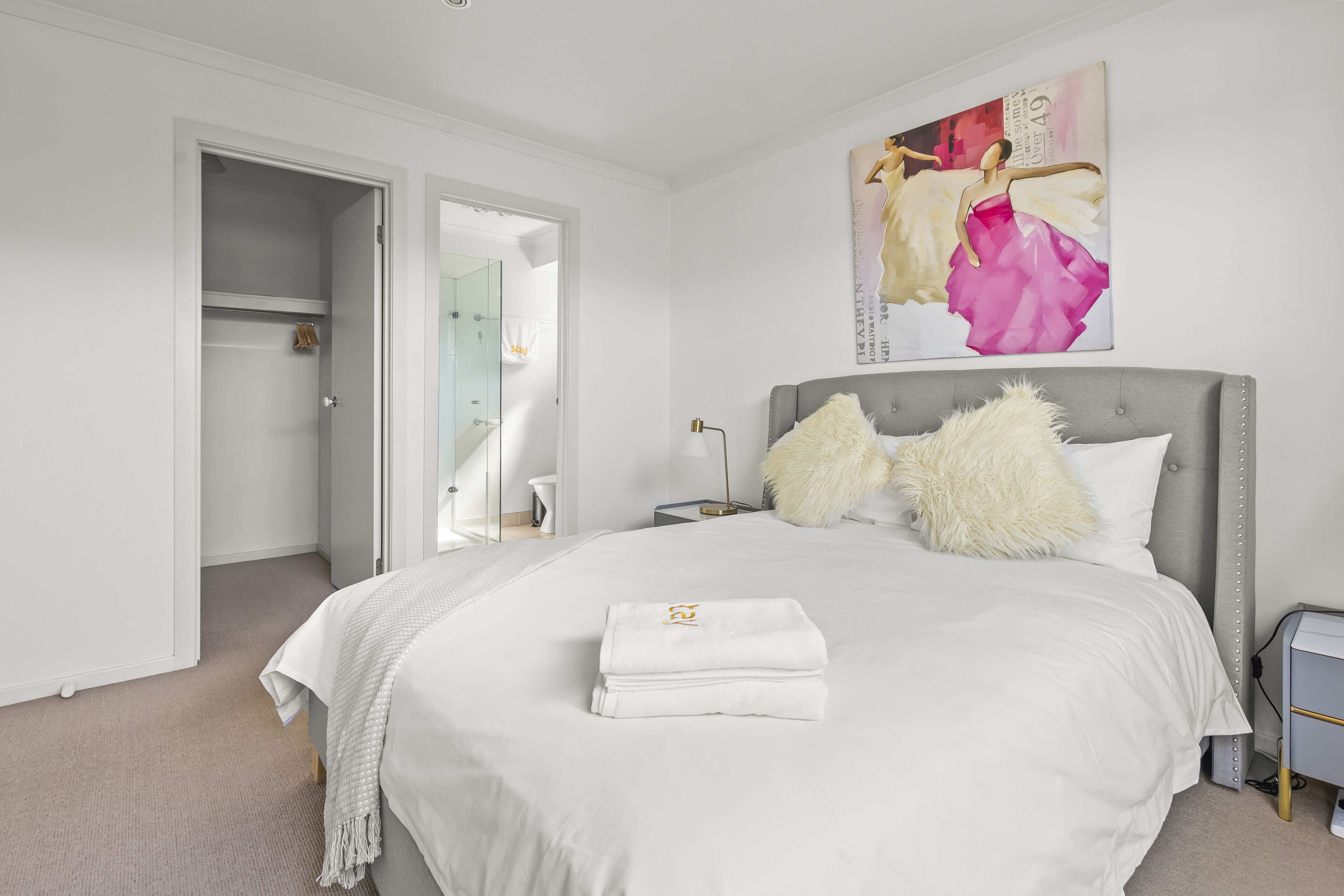 A modern bedroom with a large bed, a nightstand, a lamp, and a painting on the wall.