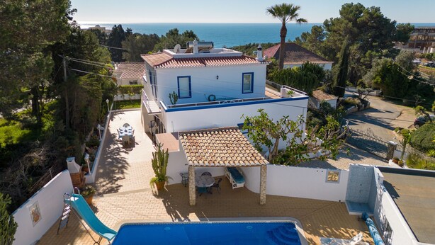 A two-story house with a swimming pool, a patio with a table and chairs, and a view of the sea.