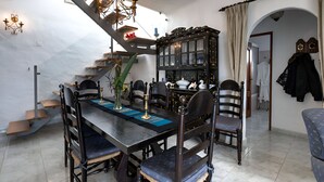 A dining area with a long table, chairs, a chandelier, a staircase, and a bookshelf.