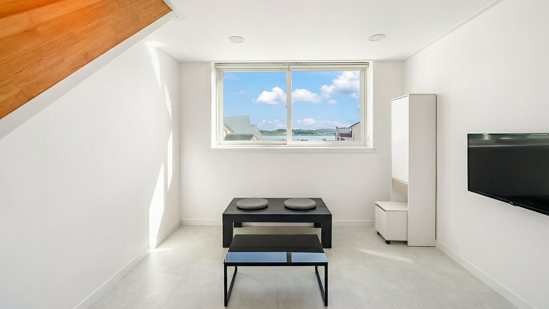 A modern, minimalistic room with a large window, a flat-screen TV, and a small table with cushions.