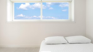 Ein Zimmer mit einem Bett, weißer Bettwäsche und einem Fenster mit Blick auf einen klaren Himmel.