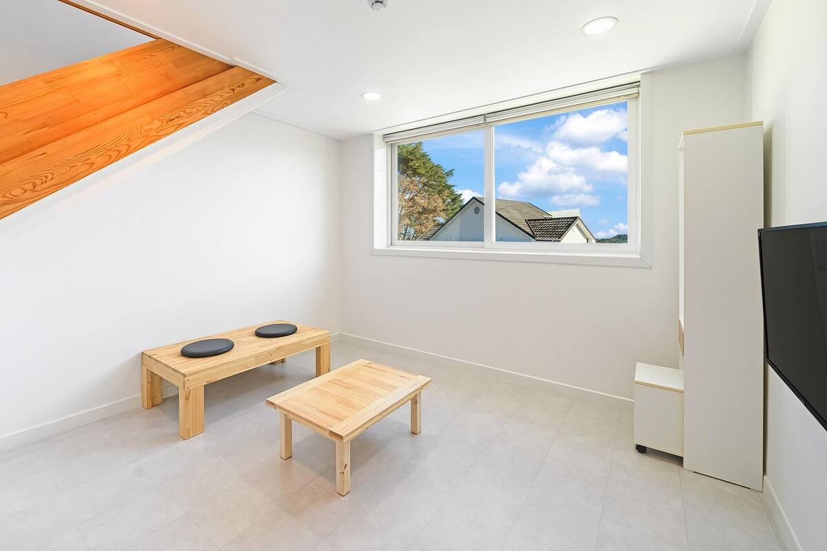 A small, modern kitchen with a built-in refrigerator, a window with a view of houses, and a wooden bench with two black stools.