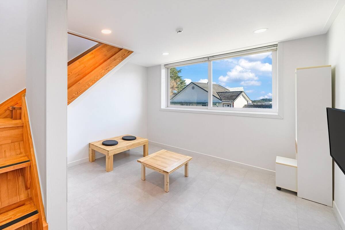 A modern apartment with a wooden staircase, a small table with two cushions, a window with a view of houses, and a flat-screen TV.
