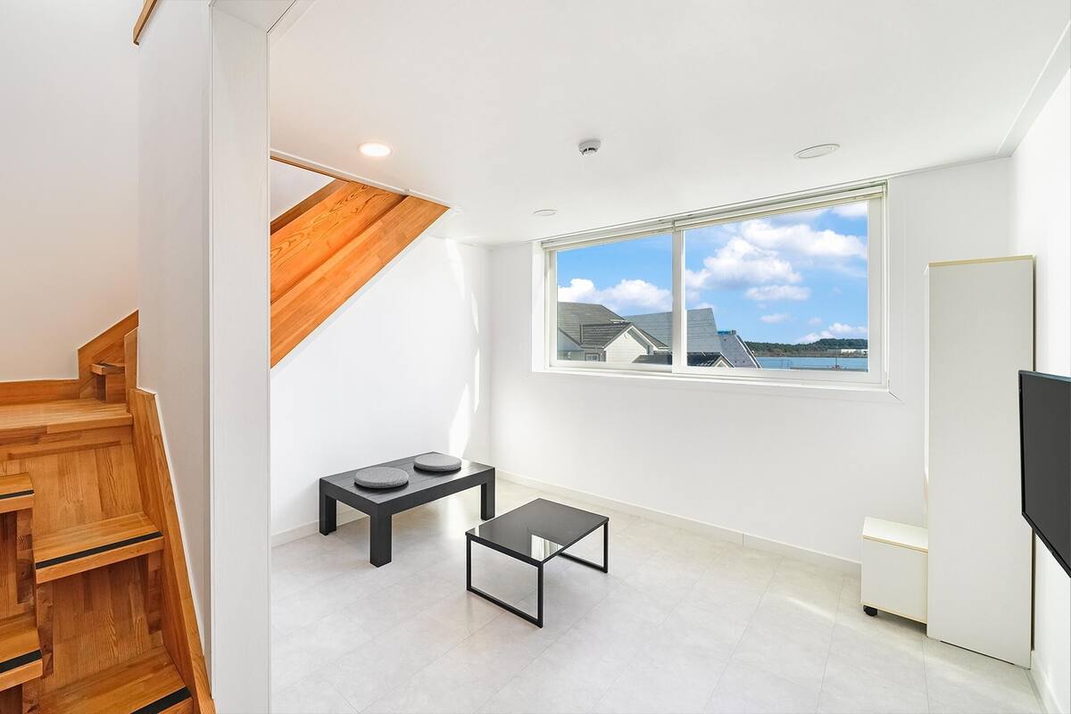 A modern apartment with a wooden staircase, a small table with two cushions, a TV, and a large window offering a view of houses and a body of water.
