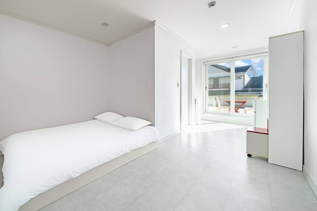 A modern, minimalist bedroom with a large bed, a sliding glass door leading to a balcony, and a clean, white interior.